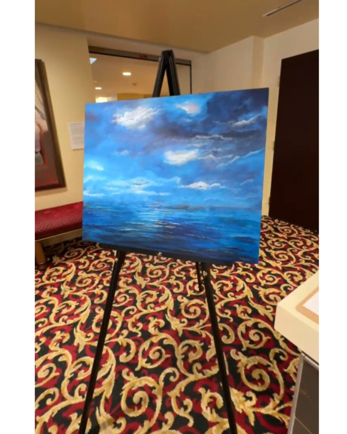 "Hope Between Storms,” an acrylic painting by Kellyanne featuring sunlight breaking through dramatic blue storm clouds over a calm ocean, displayed on an easel at a charity gala.